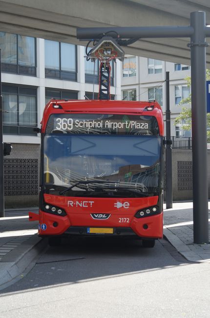 A red electric bus parked at a dedicated bus stop in an urban area, with a modern glass-fronted building in the background. The bus displays route information for line 369 servicing Schiphol Airport and Plaza, with clear windows showing the interior. The bus appears clean and well-maintained, with a smooth, shiny exterior and a modern design. Overhead, part of the bus shelter's roof and a pole supporting it are visible, with adequate natural lighting illuminating the scene. This image reflects surface and vehicle cleaning standards in a commercial setting, highlighting the importance of proper maintenance and cleanliness in transportation environments, as emphasized by Carpet Cleaners W8 for comprehensive cleaning and sanitisation services.