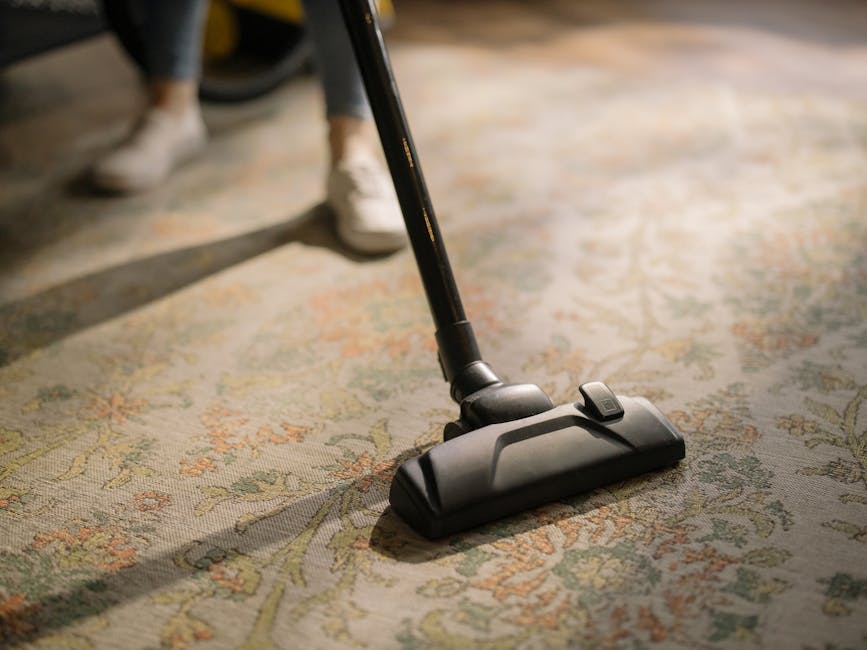 Close-up view of a person vacuuming a traditional patterned area rug with a black handheld vacuum cleaner, the rug featuring intricate floral designs in muted green, beige, and orange tones. The environment is well-lit with natural light, highlighting the cleanliness and condition of the rug. In the background, a person wearing white shoes and dark trousers is visible, suggesting a domestic setting. The scene illustrates surface cleaning and deep cleaning practices as part of regular home maintenance, with Carpet Cleaners W8 providing professional sanitisation and domestic cleaning services in the W8 area.