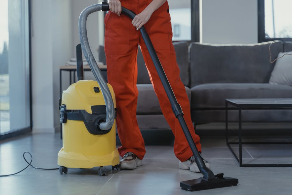 A professional cleaner wearing orange overalls is using a yellow vacuum cleaner with a black hose and a metal wand to perform surface cleaning on a tiled floor in a modern living room. The room features large windows allowing natural light, a dark gray fabric sofa with cushions, and a black metal coffee table. The vacuum cleaner is positioned on the tiled floor, which appears clean and dry, with the cleaner focused on removing dust and debris. The setting highlights hygienic maintenance within a domestic space, indicative of deep cleaning services provided by Carpet Cleaners W8 for residential environments, specifically around Holland Park Avenue in W8.