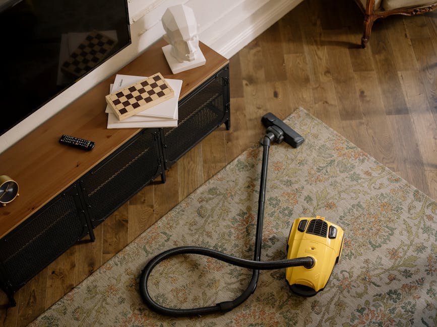 A yellow canister vacuum cleaner with a black hose and a metal extension tube is positioned on a decorative beige and green patterned rug in a living room. The room features wooden flooring and a low black metal console table against a white wall, which holds a white bust sculpture, a remote control, a stack of papers and a chessboard. The lighting appears natural, highlighting the clean, polished wooden floor and well-maintained surfaces, reflecting the professional deep cleaning services provided by Carpet Cleaners W8 for surface cleaning and sanitisation in residential environments.