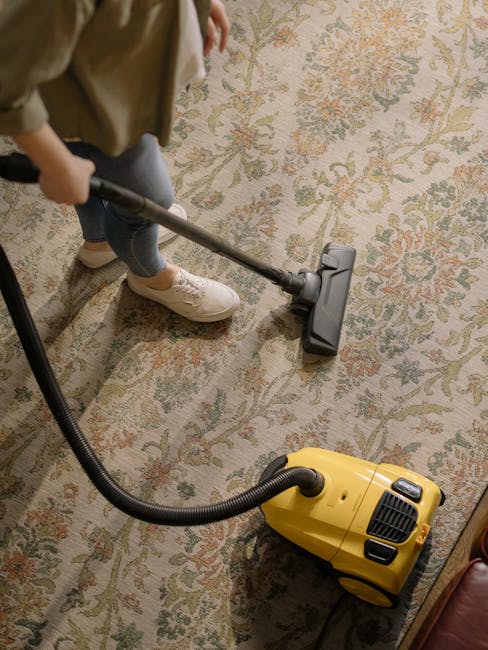 A professional cleaner wearing orange overalls is using a yellow vacuum cleaner with a black hose and a metal wand to perform surface cleaning on a tiled floor in a modern living room. The room features large windows allowing natural light, a dark gray fabric sofa with cushions, and a black metal coffee table. The vacuum cleaner is positioned on the tiled floor, which appears clean and dry, with the cleaner focused on removing dust and debris. The setting highlights hygienic maintenance within a domestic space, indicative of deep cleaning services provided by Carpet Cleaners W8 for residential environments, specifically around Holland Park Avenue in W8.