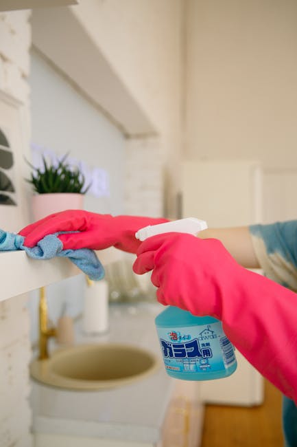 A person wearing pink rubber gloves is holding a white spray bottle with blue liquid cleaner, preparing to disinfect a white kitchen windowsill. The windowsill features a small potted plant with green foliage in a pink pot, set against a white painted brick wall and a light-colored wall in the background. The surface appears clean, dust-free, and well-maintained, with natural lighting illuminating the area, emphasizing a tidy and hygienic environment suitable for detailed surface cleaning as part of an end of tenancy cleaning checklist for landlords, W8, by Carpet Cleaners W8.
