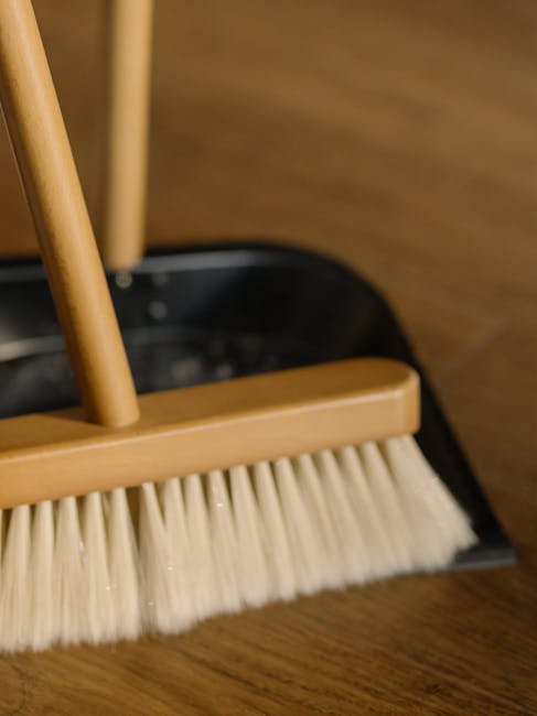 Close-up of a dustpan and cleaning brush with cream-colored bristles, positioned on a wooden floor in a room with warm lighting. The brush appears flat and wide, suitable for surface cleaning, and is part of a cleaning tool set used for dusting and sweeping hard surfaces. The wooden floor has a smooth, polished finish, indicating recent cleaning or maintenance. In the background, a portion of a wooden chair leg is visible, suggesting the setting is a living or dining area. The image highlights the tools and environment associated with domestic surface cleaning, reflecting the thorough cleaning processes recommended for end-of-tenancy preparations, as seen in the Holland Park end of tenancy cleaning checklist for landlords provided by Carpet Cleaners W8.
