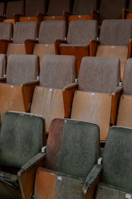 An array of aged auditorium seats arranged in rows, featuring wooden armrests and predominantly upholstered in green and beige fabric, with some showing visible wear and dirt. The seats are set on a metal framework, and the overall scene suggests a need for deep cleaning and maintenance. The lighting is dim, emphasizing the dust and grime accumulated on the fabric and surfaces. This image exemplifies the importance of comprehensive surface cleaning and sanitisation for occupied or neglected public venues, as provided by Carpet Cleaners W8, as showcased in the Ilchester Place upholstery cleaning before and after case study, W8.