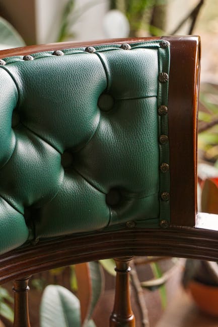Close-up of a vintage wooden armchair with a green leather upholstered backrest and seat, featuring button-tufted detailing. The leather surface appears clean and polished, reflecting light to showcase its smooth, shiny finish. The wooden frame and armrests are dark-stained, contrasting with the vibrant green upholstery. The background includes a blurred indoor setting with houseplants and natural light, emphasizing a tidy and well-maintained environment. This image illustrates the results of upholstery cleaning, highlighting the furniture's overall cleanliness and shine as part of a deep cleaning or sanitisation process conducted by Carpet Cleaners W8 in a residential space.